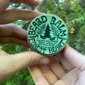 Peut inclure: Une boîte ronde et verte de baume à barbe. La boîte présente les mots "BEARD BALM" dans une police incurvée, avec un graphique de forêt et les mots "FOREST HEART". Les ingrédients comprennent de l'épinette noire, du cèdre et des huiles essentielles de citron.