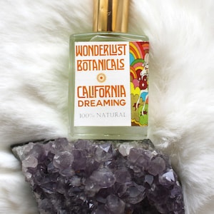 May include: A clear glass perfume bottle with a gold cap, labeled "Wonderlust Botanicals California Dreaming 100% Natural." The bottle sits atop a cluster of purple amethyst crystals, against a white, fluffy background.