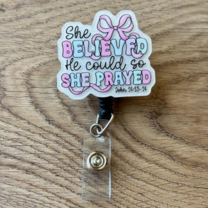 May include: A badge reel featuring a white acrylic topper with a pink bow graphic and the text She BELIEVED He could so SHE PRAYED John 14:13-14 in pink and blue bubble letters. The reel has a black plastic housing, a retractable cord, and a clear strap.