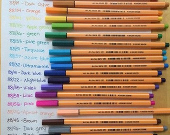 Puede incluir: Un conjunto de 18 marcadores de colores con una variedad de colores, incluyendo rojo, naranja, amarillo, verde, azul, morado, rosa, marrón, gris y negro. Los marcadores están etiquetados con su color y número. Los marcadores están dispuestos en una fila sobre una superficie blanca.