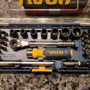 May include: A blue plastic case containing a set of tools, including sockets, a ratchet, and a screwdriver handle. The brand name "VACO" is visible in yellow and black. The case also includes the text "SAE: VAC3320 Metric: VAC3321".