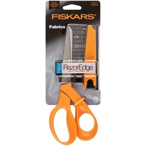 May include: Orange Fiskars RazorEdge fabric shears with a black blade and a clear plastic sheath. The packaging says "New RazorEdge premium fabric shears" and lists the fabrics the shears can cut: suede, corduroy, silk, oilcloth, damask, denim, terry cloth, polyester, and crepe.