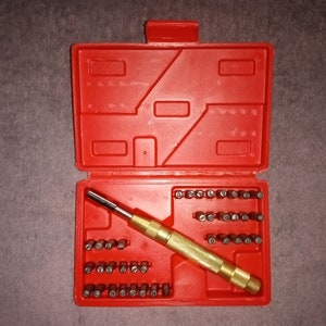May include: A red plastic tool case with a hinged lid, open to reveal a set of small screwdriver bits and a gold-colored handle. The bits are arranged in rows, and the handle is in the center.