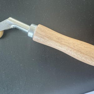 May include: A metal and wood tool with a small, round, brass-coloured wheel. The tool has a wooden handle and a silver-coloured metal frame. The wheel is approximately 2.5 cm in diameter. The tool is designed for various crafting or construction tasks.