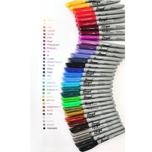 May include: A collection of colorful permanent markers arranged in a curved line. The markers have gray barrels and various colored tips, including yellow, red, purple, blue, and green. The text "Pragel" is printed on the markers.