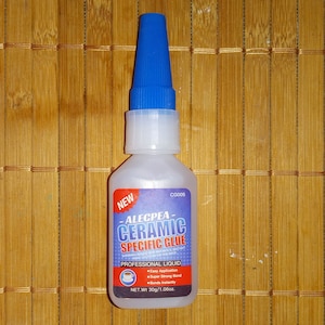 Può includere: Una bottiglia di plastica trasparente di Alecpea Ceramic Specific Glue con un tappo blu. La bottiglia è etichettata "NEW" e ha un'etichetta bianca con testo blu. La bottiglia è appoggiata su una superficie di bambù marrone.