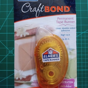 May include: A yellow and clear plastic tape dispenser with the brand name "Elmer's" and the text "CraftBOND Permanent Tape Runner" on the packaging. The dispenser has a clear window showing the tape inside. The packaging also includes the text "clear, double-sided adhesive, high tack, 6.25 ft, .31 in x 26.25 ft (8 mm x 8 m), .31 in actual width".