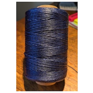 May include: A spool of dark blue thread. The thread is tightly wound around a wooden core, creating a cylindrical shape. The thread has a slightly textured appearance, and the color is a deep, rich blue.
