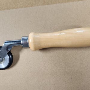 Puede incluir: Una herramienta manual con un mango de madera clara y una rueda de metal. La rueda es negra y plateada. La herramienta se utiliza probablemente para alisar o prensar materiales.