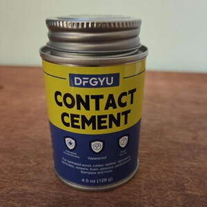 Może przedstawiać: Cylindryczny pojemnik z klejem kontaktowym DFGYU. Pojemnik ma srebrną pokrywę i żółto-niebieską etykietę. Etykieta zawiera słowa "CONTACT CEMENT" dużymi, pogrubionymi literami. Pojemnik zawiera 128 g kleju.