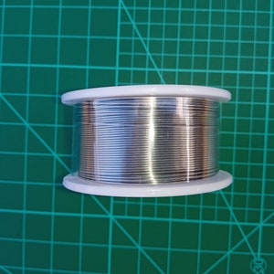 Puede incluir: Una bobina de alambre de soldadura de plata en un contenedor de plástico blanco. El contenedor tiene una tapa de plástico transparente.