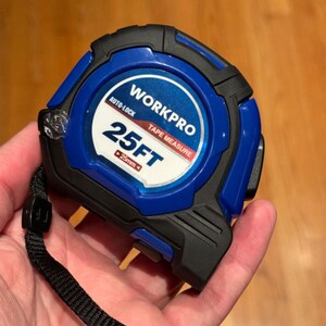 Puede incluir: Una cinta métrica azul y negra con una etiqueta blanca que dice "WORKPRO TAPE MEASURE 25FT AUTO-LOCK 25mm".