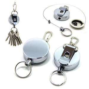May include: Three retractable keychains with a silver finish. Each keychain has a round metal housing with a spring-loaded retractable cord. The keychains are attached to a metal clip and a ring.