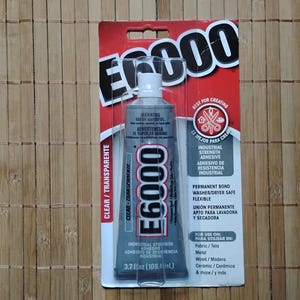 May include: E6000 industrial strength adhesive in a clear tube, packaged on a red and white card. The label indicates it is permanent bond, washer/dryer safe, and flexible. The tube contains 3.7 fl oz (109.4 ml).