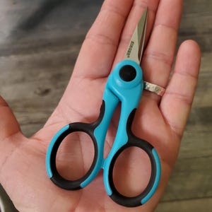 May include: A pair of small, blue scissors with black handles. The scissors have a silver blade with the word "Bircher" printed on it.