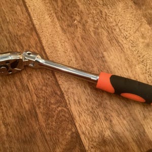 Könnte beinhalten: Eine verchromte Knarrenschlüssel mit einem schwarzen und orangefarbenen Gummigriff. Dieses Werkzeug wird zum Anziehen oder Lösen von Muttern und Bolzen verwendet. Der Griff ist für Komfort und Kontrolle konzipiert. Ca. 20 cm lang.