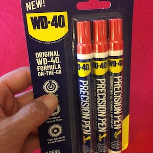 Może przedstawiać: Opakowanie z trzema czerwonymi długopisami precyzyjnymi WD-40. Długopisy są oznaczone "Oryginalna formuła WD-40 On-The-Go" i zapakowane na niebieskim i żółtym tle. Opakowanie zawiera napis "Nowość!" i "3 długopisy 0,3 fl. oz. (9 ml) każdy".