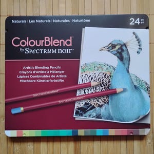 May include: A box set of 24 ColourBlend artist's blending pencils by Spectrum Noir. The box features a peacock illustration and the text "ColourBlend by Spectrum Noir". Two pencils are visible, one with a blue tip. The box is on a bamboo surface.
