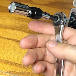 Op de afbeelding: Een verchroomde ratelsleutel met een zwarte handgreep en een dop bevestigd. De dop is zilverkleurig en heeft een zeshoekige opening. De tool is ontworpen voor het aandraaien of losdraaien van bouten en moeren.