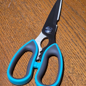 May include: Kitchen shears with black blades and a silver hinge. The handles are gray with teal accents. The shears have a serrated section and a bottle opener. The shears are on a wooden surface.