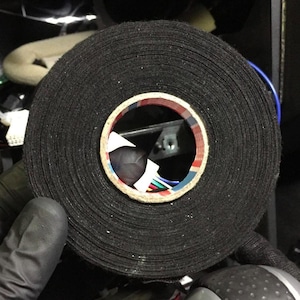 Könnte beinhalten: Eine große Rolle schwarzes Isolierband. Das Band ist eng gewickelt und zeigt die Materialschichten. Die Mitte der Rolle zeigt einen Pappkern mit farbigen Drähten im Inneren. Das Band wird von einer behandschuhten Hand gehalten.
