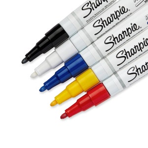 Könnte beinhalten: Ein Satz von sechs Sharpie Permanentmarkern in Schwarz, Weiß, Blau, Gelb, Rot und Silber. Die Marker sind fächerförmig angeordnet, wobei die Spitzen zum Betrachter zeigen. Die Marker sind alle mit dem Markennamen Sharpie und Gebrauchsanweisungen versehen.