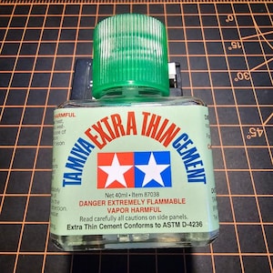 May include: A bottle of Tamiya Extra Thin Cement, a model-making adhesive. The bottle has a green cap and a label with red, white, and blue stars. The label also includes the text "DANGER EXTREMELY FLAMMABLE" and "VAPOR HARMFUL". The bottle contains 40ml.