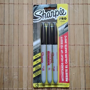 May include: A pack of three Sharpie Pro Industrial permanent markers. The markers have black caps and grey barrels. The packaging is yellow and black, with text indicating the markers withstand heat up to 260°C.