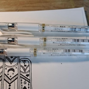 Könnte beinhalten: Drei weiße Gel-Stifte mit schwarzer Tinte. Die Stifte sind mit "Weiß" beschriftet und haben eine 1,0 mm Kugelspitze. Die Stifte sind für die Verwendung auf Illustrationen und schwarzem Papier konzipiert.