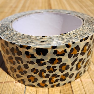 May include: A roll of leopard print duct tape. The tape features a tan background with brown and black spots. The roll is sitting on a wooden surface.