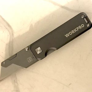Puede incluir: Un cúter WORKPRO gris con una cuchilla plateada. El cuchillo está abierto, revelando la afilada cuchilla. El mango es rectangular con bordes redondeados y tiene un botón y un pequeño logotipo. El cuchillo está sobre una superficie clara.