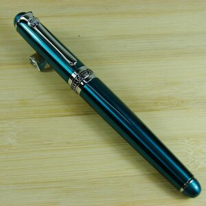 May include: A teal blue fountain pen with silver accents and a decorative design. The pen has a cap and a clip.