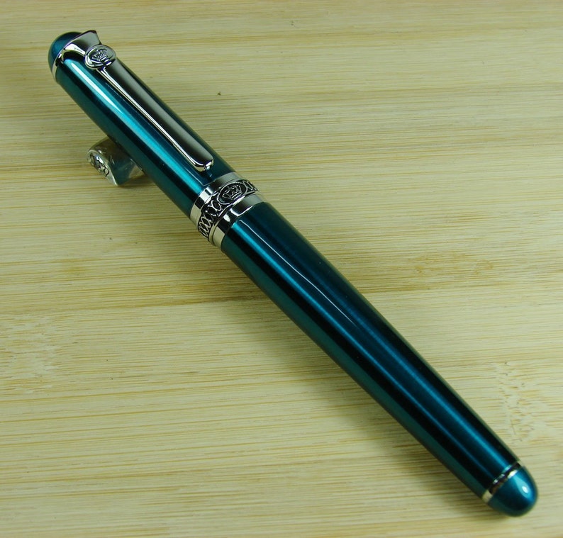 May include: A teal blue fountain pen with silver accents and a decorative design. The pen has a cap and a clip.