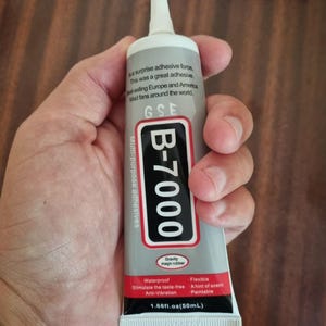 以下が含まれることがあります： 白い先端と灰色の本体が付いた、B-7000多目的接着剤のチューブ。 チューブには「B-7000」と「50ml」を含む黒と赤のテキストがあります。 テキストはまた、防水性と柔軟性があることにも言及しています。