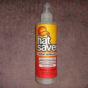 Puede incluir: Una botella de plástico transparente de Hat Saver Repelente de Sudor. La botella es amarilla y naranja con un logotipo de gorra de béisbol roja. La etiqueta dice "Hat Saver Repelente de Sudor", "Repels Sweat & Stains", "Keep Hats Looking New", "Protect All Hats", "WARNING: FLAMMABLE", "Read precautions on back", "NET 8 FL OZ. (237 ml)"