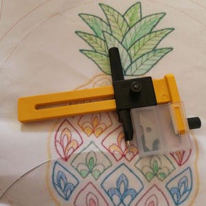 May include: A yellow and black Olfa rotary cutter with a clear blade cover rests on a paper with a pineapple design. The pineapple is drawn with green leaves and colorful, patterned segments.