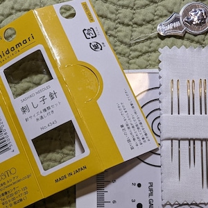 May include: A yellow and white package of sashiko needles labeled "Hidamari Sashiko" with the text "刺し子針" (Sashiko needles) and "No. 4345". The package is sitting on a green fabric surface. A small white package of needles is also visible in the image.