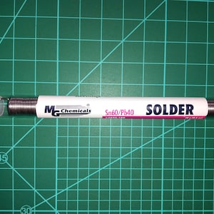 Puede incluir: Un tubo transparente de soldadura con una etiqueta blanca con el texto "MG Chemicals" y "SOLDER". La soldadura es una bobina de alambre plateado. El tubo está sobre una esterilla de corte verde.