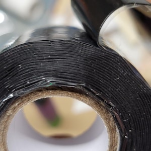 May include: Close-up of a roll of black electrical tape. The tape is tightly wound, revealing the layers of the adhesive material. The end of the roll is partially peeled back, showing the shiny, black surface.