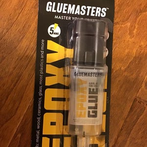May include: A clear plastic bottle of GlueMasters Epoxy Glue with a black cap. The bottle is labeled with the text "GlueMasters Epoxy Glue" and "0.51 fl. oz. (12ml + 12ml)". The packaging is yellow and black and says "Bonds: metal, wood, ceramics, glass, most plastics and more".