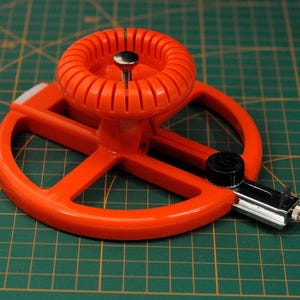 May include: An orange circle cutter tool with a central adjustment knob and a silver blade housing. The tool is set against a green grid background, suggesting precision and measurement.