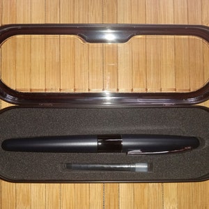 May include: A black fountain pen with a silver trim and a black ink cartridge in a black metal case with a clear plastic lid.