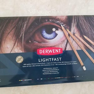Op de afbeelding: Een rechthoekige doos met Derwent Lightfast oliegebaseerde kleurpotloden. De doos heeft een oogafbeelding en de tekst "LIGHTFAST" en "36". Er liggen verschillende potloden bovenop de doos.