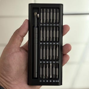 Puede incluir: Un juego de destornilladores negro con múltiples puntas, sostenido en una mano. El juego incluye un mango de destornillador y varias puntas para diferentes tipos de tornillos. Las puntas están cuidadosamente organizadas en una caja negra.