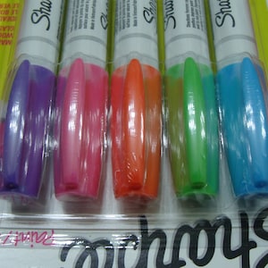 Könnte beinhalten: Eine Packung mit sechs Sharpie-Permanentmarkern in verschiedenen Farben, darunter Lila, Rosa, Orange, Grün und Blau. Die Marker sind in einem durchsichtigen Kunststoffgehäuse verpackt, auf dem das Sharpie-Logo aufgedruckt ist.