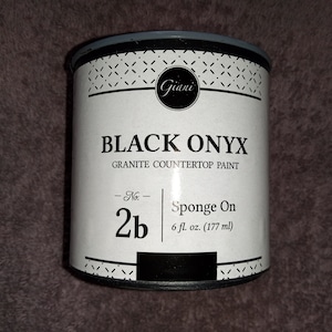 Puede incluir: Una lata de pintura para encimeras de granito Giani Black Onyx. La lata es blanca con texto negro y un patrón blanco y negro. La lata está etiquetada "No. 2b Sponge On 6 fl. oz. (177 ml)"