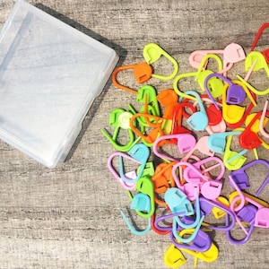 May include: A collection of colorful plastic stitch markers in various shades of pink, purple, yellow, green, orange, and blue. A clear plastic container is in the upper left corner. The stitch markers are scattered on a wooden surface.