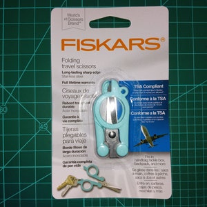 May include: A turquoise blue folding travel scissors with a TSA compliant label. The scissors are made of stainless steel and have a full lifetime warranty. The packaging includes a small image of the scissors being used to cut a piece of paper.