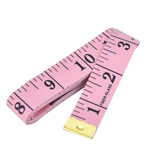 May include: A pink fiber-glass measuring tape with black markings and numbers. The tape is unrolled, showing measurements up to 10 inches. The end of the tape has a gold-colored metal tip.