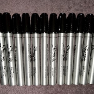 Puede incluir: Primer plano de doce marcadores permanentes Sharpie plateados con tapas negras y puntas biseladas. Los marcadores están dispuestos en fila y el texto "Sharpie" y "Punta biselada" es visible en los marcadores.